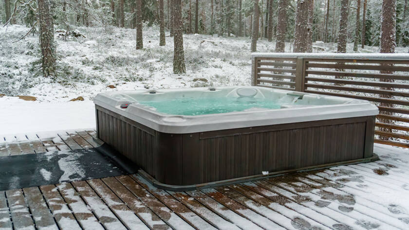 Hot tub on a wooded patio surrounded by snow with a wooden wall blocking out wind