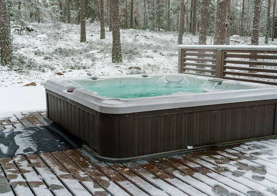 Hot tub on a wooded patio surrounded by snow with a wooden wall blocking out wind